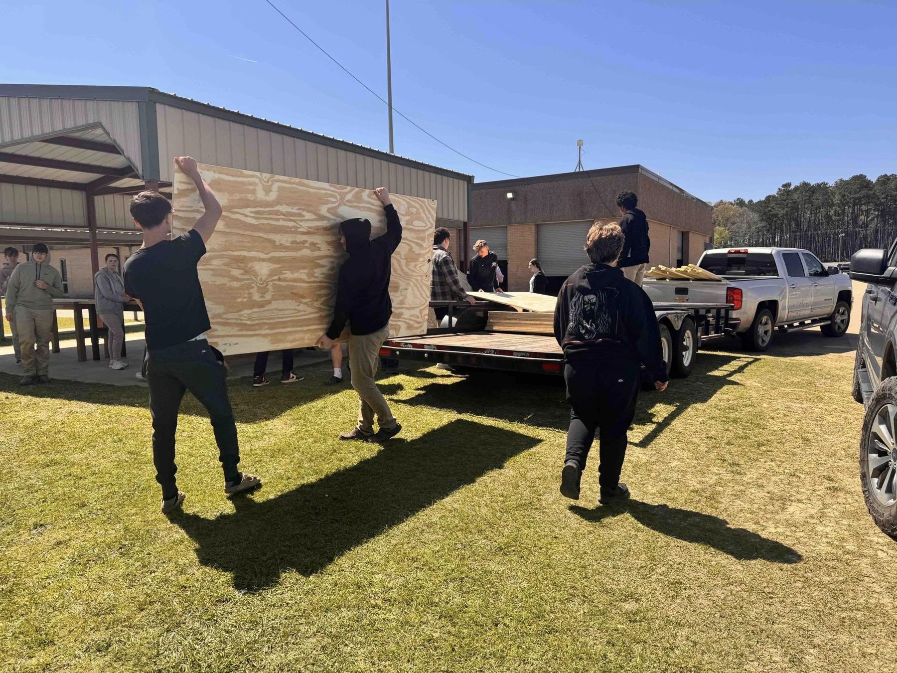 Students carrying a large plywood panel across a lawn